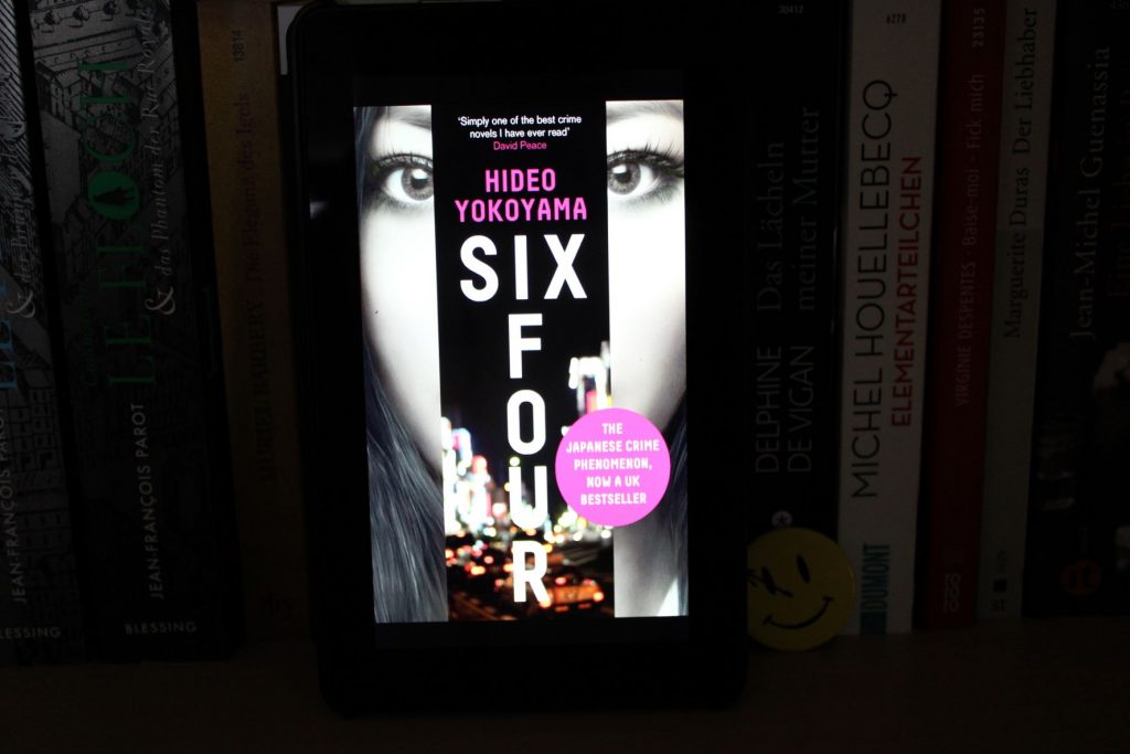 E-Book-Cover von »Six Four«. Im Hintergrund eine schwarz-weiß-Nahaufname des Kopfes einer jungen Frau mit langen schwarzen Haaren. Genau in der Mitte geht ein schmaler Streifen von oben nach unten, auf dem nächtliche Großstadtlichter zu sehen sind, dazu der Name des Autors und der Buchtitel.