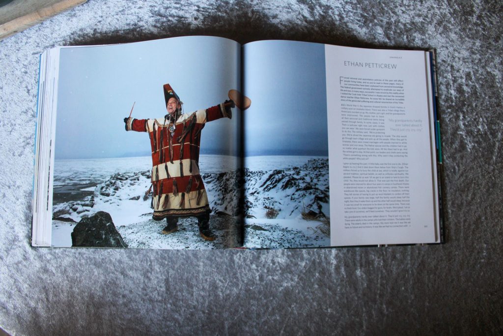 Älterer indigener Mann vor felsiger Eislandschaft in traditoneller Winterkleidung. Steht breitbeinig da, die Arme ausgebreitet, in den Händen eine kleine Trommel und einen Stock.
