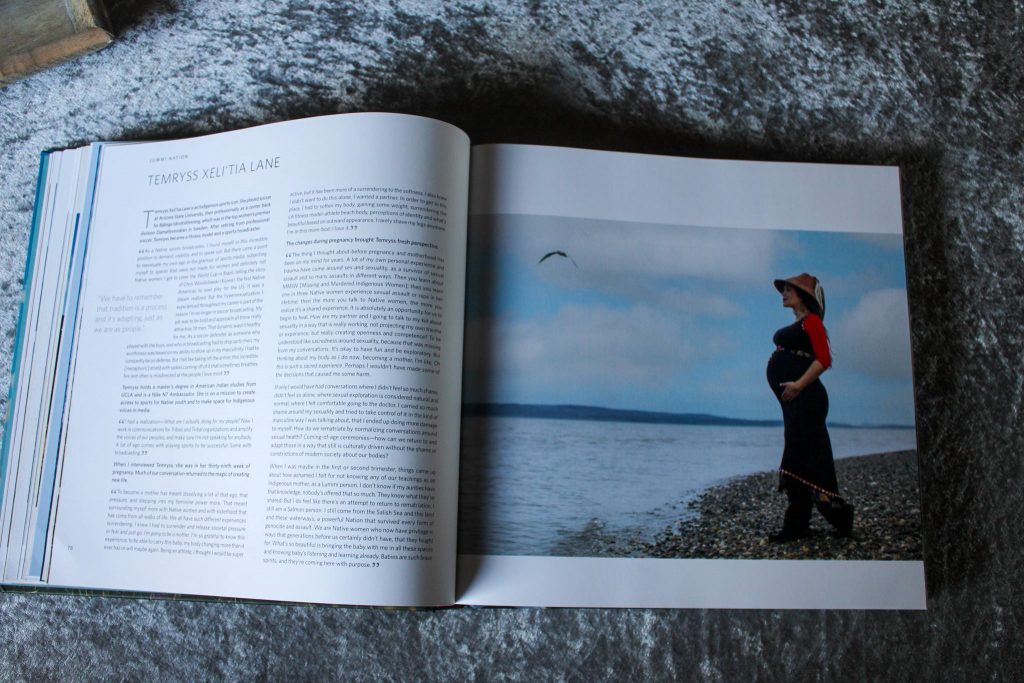 Hochschwangere indigene Frau in langem schwarzen Klein mit roten Armen. Auf dem Kopf ein Strohhut, die Hände auf den Buch gelegt, an einem Kieselsteinstrand vor einem See stehend.. Am Himmel fliegt ein größerer Vogel