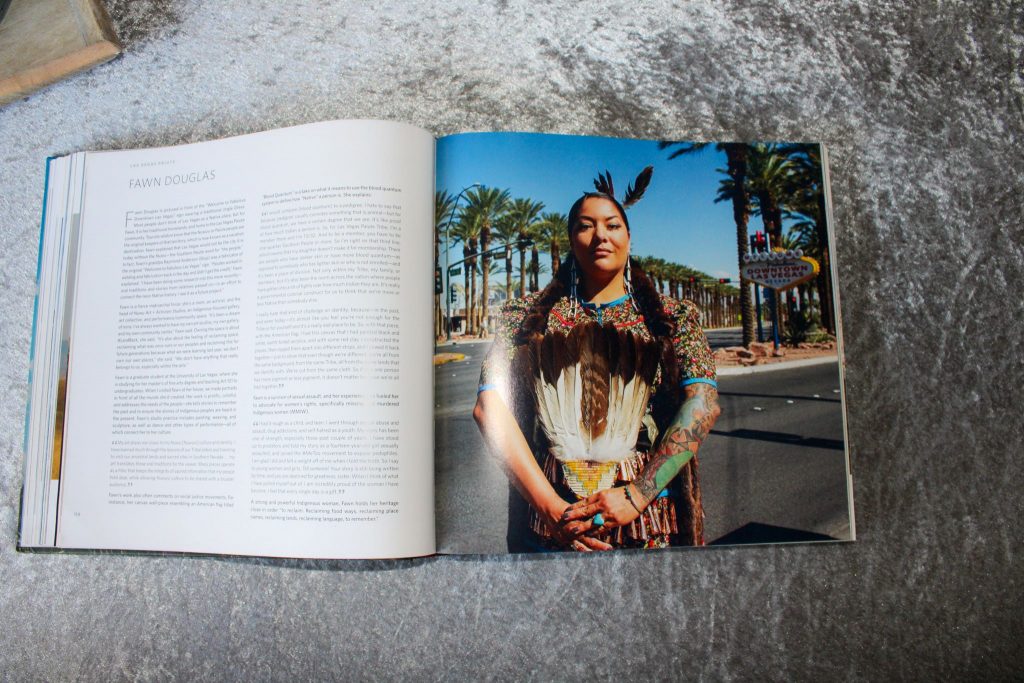 Indigene Frau mit Federschmuck und bunten Kleid an einer von Palmen gesäumten Straße, den linken Arm stark tätowiert.