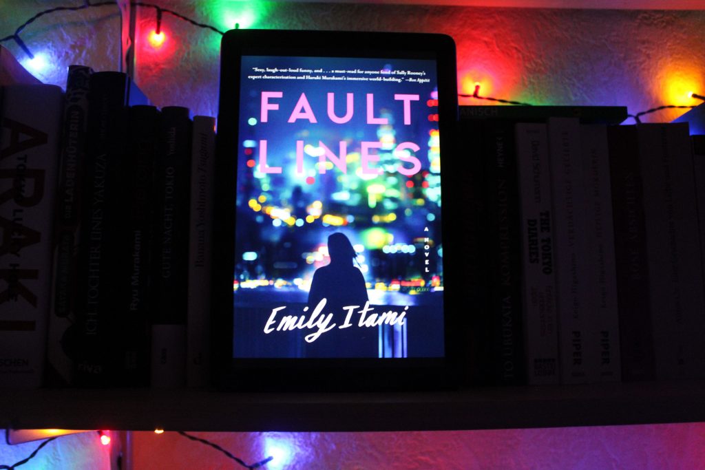 E-Book-Cover von Emily Itamis »Fault Lines« auf einem Tablet, das die schwarze Silhouette einer Frau von hinten vor der verschwommenen nächtlichen Ansicht Tokios zeigt.