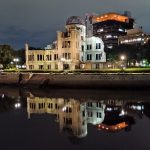Das Friedensdenkmal in Hiroshima. Die ausgebombte Ruine eines mehrstöckigen älteren Gebäudes aus Stein mit Kuppeldach, hier bei Nacht und angeleuchtet von der anderen Flussseite von leicht rechts fotografiert. Das Gebäude spiegelt sich im Fluss.