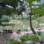 Ein japanischer Garten mit Teich in der MItte, einer überdachten Sitzbank am Teich und dahinter Rahmen zwei Bäume das Bild ein.