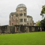 Das Friedensdenkmal in Hiroshima. Die ausgebombte Ruine eines mehrstöckigen älteren Gebäudes aus Stein mit Kuppeldach, hier am Tag aus der Nähe fotografiert. Davor eine Mauer, eine grüne Wiese und rechts im Bild ein Baum.