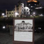 Das Friedensdenkmal in Hiroshima. Die ausgebombte Ruine eines mehrstöckigen älteren Gebäudes aus Stein mit Kuppeldach, hier bei Nacht und angeleuchtet von der anderen Flussseite mittig fotografiert. Das Gebäude spiegelt sich im Fluss. Auf unserer Flussseite steht ein Hinweisschild mit Foto und japanischen Schriftzeichen.