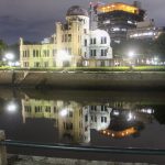 Das Friedensdenkmal in Hiroshima. Die ausgebombte Ruine eines mehrstöckigen älteren Gebäudes aus Stein mit Kuppeldach, hier bei Nacht und angeleuchtet von der anderen Flussseite mittig fotografiert. Das Gebäude spiegelt sich im Fluss.